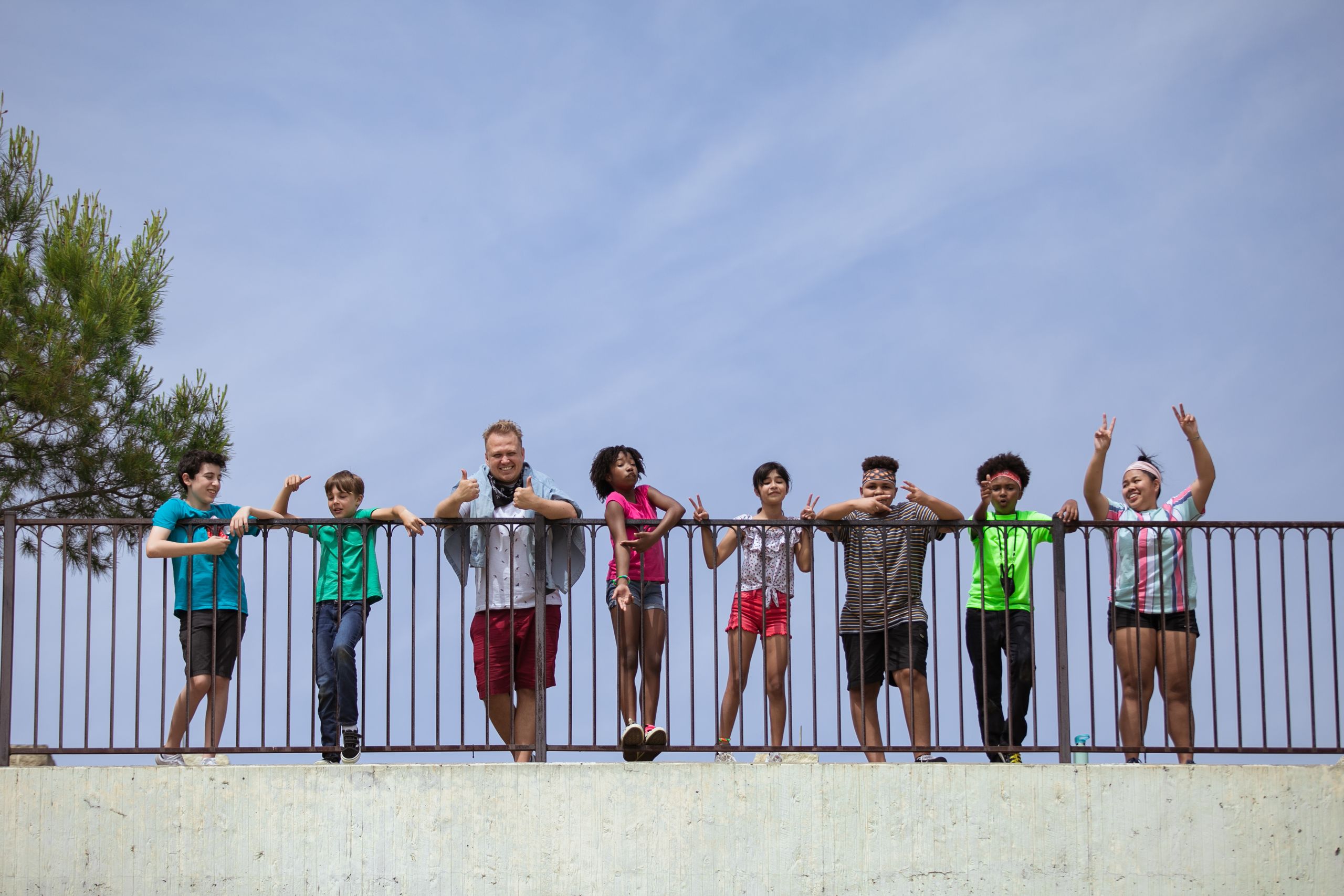 A diverse group of friends standing on a metal fence outdoors on a sunny day, expressing joy.